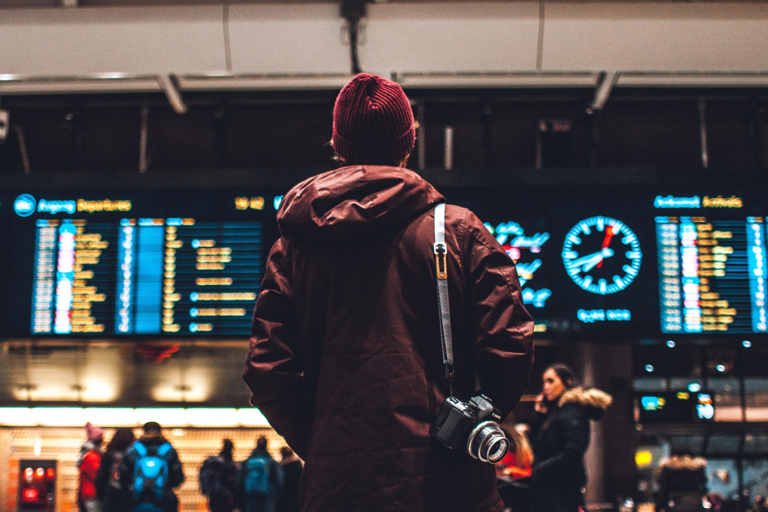 Man stands with his back to the camera, he has a camera slung over his shoulder. He is looking at a departures and arrivals screen.